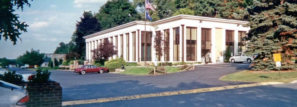 A two-story white building with windows reaching from the ground to the roof. A parking lot with retro cars.